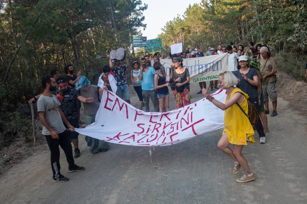 Protesters on a dirt road. They hold a banner saying "emperyalist sirket kazdagini terket". They are protesting the Alamos Gold mine near Kirazlı.