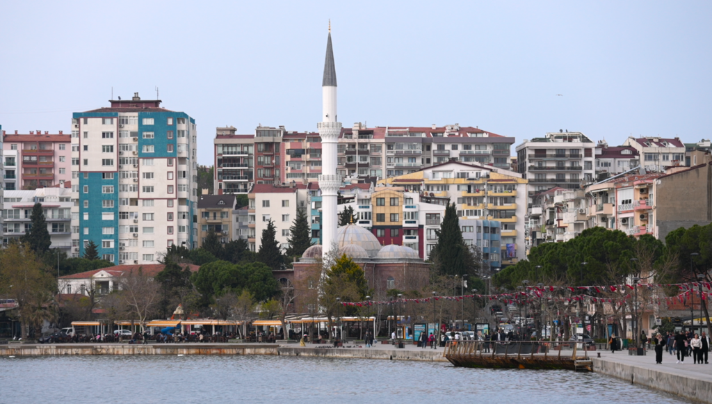 Sea front of Çanakkale, Türkiye, with a white mosque, apartment buildings and people walking along the water side.